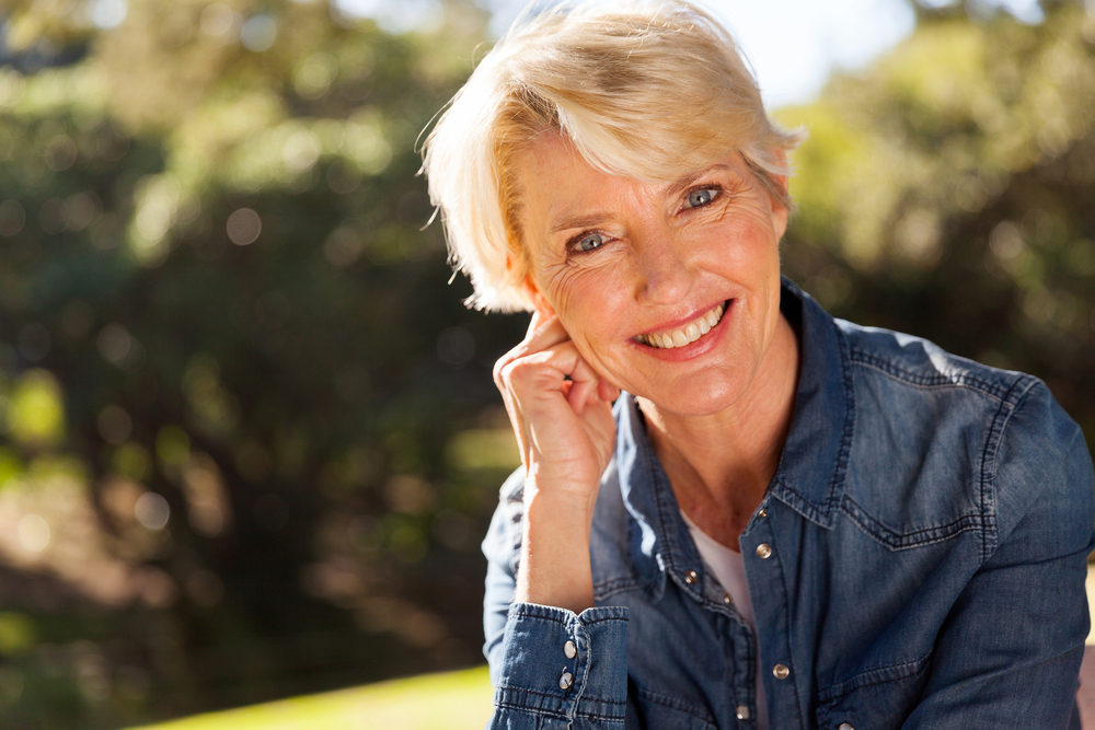 Mature woman with short blonde hair wearing a denim buttonup shirt and smiling outdoors